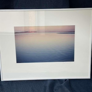 Puede incluir: Fotografía enmarcada de un paisaje marino sereno con una paleta de colores suaves y pastel. El agua presenta un degradado de azules, y el cielo muestra tonos rosados y anaranjados. El marco blanco añade una estética limpia y moderna a la obra.