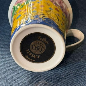 Puede incluir: Una taza de porcelana con un vibrante patrón floral en amarillo, azul y rosa. La base muestra una etiqueta circular negra con el texto Rosenthal, Versace y Primavera, junto con un logotipo de cabeza de Medusa y un borde dorado.
