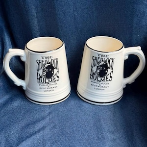 Puede incluir: Dos tazas de cerámica color hueso con un gráfico negro de una silueta con gorra de cazador y fumando en pipa. El texto dice THE SHERLOCK HOLMES PUBLIC HOUSE & RESTAURANT LONDON. Cada taza tiene un borde negro y una franja negra en la base.