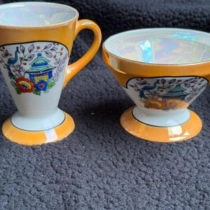 Puede incluir: Una jarra de leche y un azucarero de cerámica a juego con borde y base de color naranja vibrante e interior iridiscente. Ambas piezas están decoradas con un estampado gráfico colorido de una pagoda, un pavo real y flores sobre fondo blanco.