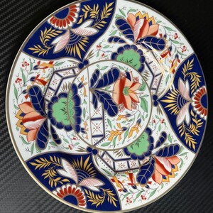 Puede incluir: Un plato redondo de cerámica con un patrón complejo de motivos florales en azul oscuro, naranja, verde y dorado. El diseño incluye flores estilizadas, hojas y formas geométricas dispuestas simétricamente con un borde dorado.