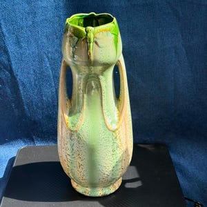 May include: A ceramic vase featuring a light green glaze with darker green drips near the rim and brown speckled accents. The vase has a tall, slender body with two integrated side handles that extend from the neck to the mid-section of the vessel.