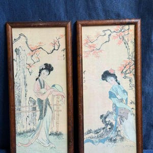 Puede incluir: Dos grabados enmarcados de estilo asiático. Cada grabado presenta a una mujer con vestimenta tradicional, sobre un fondo de árboles y follaje. Los marcos son de madera oscura. La obra de arte está en colores pastel suaves.