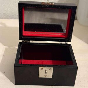 Puede incluir: Caja de joyería negra abierta con interior de terciopelo rojo. La caja tiene una tapa con bisagras con una parte superior espejada y un cierre plateado en forma de engranaje. Una cerradura plateada está en la parte delantera.
