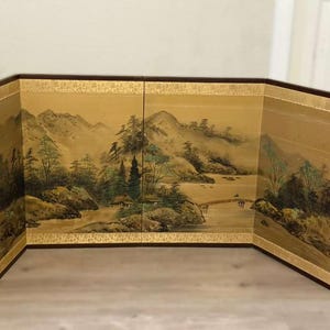 Puede incluir: Un biombo con una pintura de paisaje. La obra representa montañas, árboles y un río con un puente. La pantalla tiene un borde dorado y un marco marrón oscuro. La obra de arte está en tonos apagados.