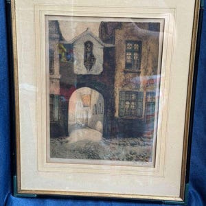 May include: A framed etching depicting a European street scene with a stone archway, old buildings with shuttered windows, and a small statue in a niche above the arch. The artwork is encased in a gold and black frame with a cream-colored mat board.