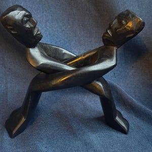 Puede incluir: Una escultura de madera de color marrón oscuro que representa dos figuras estilizadas bloqueadas en una pose de lucha o agarre. Las figuras están talladas con líneas suaves y angulares, mostrando rasgos faciales definidos y extremidades que se cruzan en una composición dinámica.