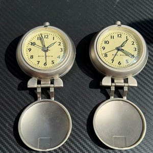 Puede incluir: Dos despertadores de viaje de metal plateado con cajas redondas y tapas con bisagras. Las esferas de color crema presentan números arábigos negros, manecillas negras y el texto "BIG BEN" impreso en negro sobre la esfera del reloj.