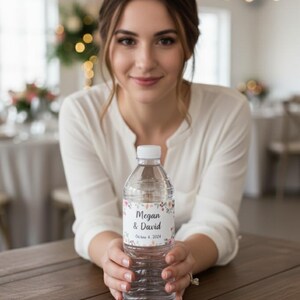 May include: Clear plastic water bottle with a white cap. The bottle has a label with the names "Megan & David" and the date "October 4, 2024" surrounded by floral designs. The bottle is held by a person.