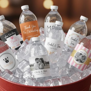 May include: Clear plastic water bottles with custom labels featuring various designs, including names, dates, and photographs, nestled in a red bucket filled with ice cubes. The labels display text such as Thank You For Celebrating, Rachel & Tom, and Olivia & Alex.