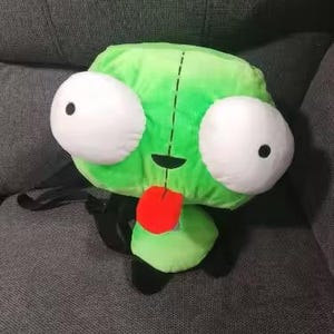 May include: A plush green character with large white eyes and black pupils, a red tongue, and a black mouth. The character has a black stitched line down the center and is sitting on a gray surface. The plush toy is a cartoon character.