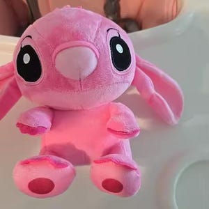 Puede incluir: Un peluche rosa de un personaje con grandes ojos negros, orejas largas y una nariz peque&ntilde;a. El juguete tiene una textura suave y difusa y est&aacute; sobre una superficie blanca. Los pies y las manos del juguete tienen acentos rosas m&aacute;s oscuros.