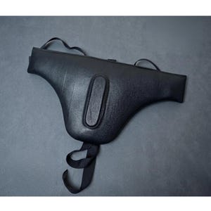 May include: A black leather hip bag with a curved shape and a central, elongated, padded detail. The bag has two straps at the top and a long strap with a loop at the bottom. The bag is shown on a gray background.