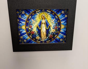 Op de afbeelding: Een rechthoekig wandkunstwerk met een afbeelding in glas-in-loodstijl van een figuur in een blauw-wit gewaad, omringd door een cirkelvormige rand van rozen, lelies en twee witte duiven, geplaatst in een massief zwart frame.