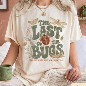May include: An ivory t-shirt featuring a graphic print with the text THE LAST OF THE BUGS and CALL ME WHEN THE BUGS DON'T DIE. The design includes illustrations of a ladybird, a dragonfly, a butterfly, a grasshopper, and various botanical elements.