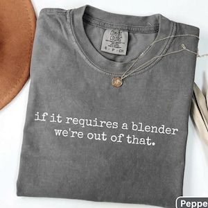 Op de afbeelding: Een antracietgrijs T-shirt met korte mouwen, een ronde hals en de witte tekst 'if it requires a blender we're out of that.' Het shirt is voorzien van een Comfort Colors-label in de nek en een verfijnde goudkleurige ketting.