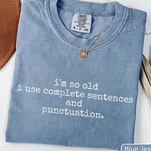Könnte beinhalten: Ein jeansblaues T-Shirt mit Rundhalsausschnitt und einem Comfort Colors Markenetikett. Auf der Vorderseite befindet sich ein weißer Aufdruck mit dem Text, i'm so old i use complete sentences and punctuation. Eine dünne goldene Kette liegt darauf.