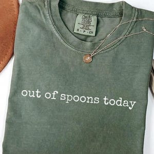 Może przedstawiać: Oliwkowozielony t-shirt z krótkim rękawem, okrągłym dekoltem i nadrukowanym na piersi białym napisem "out of spoons today". Koszulka posiada metkę marki Comfort Colors przy wewnętrznej stronie kołnierza oraz delikatny złoty naszyjnik.