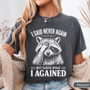Pode incluir: Uma t-shirt de manga curta cinzento-carvão com um gráfico branco de um guaxinim a cobrir os olhos com uma pata. O texto acima do guaxinim diz I SAID NEVER AGAIN e o texto abaixo diz BUT GUESS WHAT I AGAINED em letras brancas.