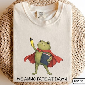 May include: An ivory t-shirt featuring a graphic of a green frog wearing a red cape, holding a yellow highlighter and a dark blue book. The text "WE ANNOTATE AT DAWN" is printed in black below the frog. The shirt has a Comfort Colors brand label.