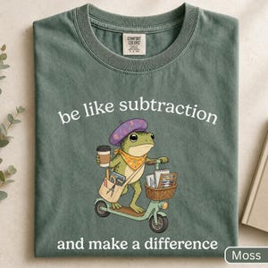 以下が含まれることがあります： モスグリーンのクルーネックTシャツ。紫のベレー帽と黄色のスカーフを身につけ、コーヒーカップを持ってスクーターに乗るカエルのグラフィックが描かれています。白い文字で「be like subtraction and make a difference」と書かれています。