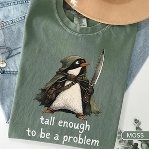 Könnte beinhalten: Ein moosgrünes T-Shirt mit einer Grafik eines Pinguins, der einen Kapuzenumhang trägt und ein Messer hält. Der Text unter dem Pinguin lautet "tall enough to be a problem". Das T-Shirt hat einen Rundhalsausschnitt und ein sichtbares Markenetikett.