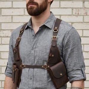 Op de afbeelding: Een bruin leren borstharnas met meerdere zakken en verstelbare riemen. Het harnas wordt gedragen over een grijs overhemd met knopen en een blauwe spijkerbroek. Het harnas heeft een robuuste, vintage uitstraling.
