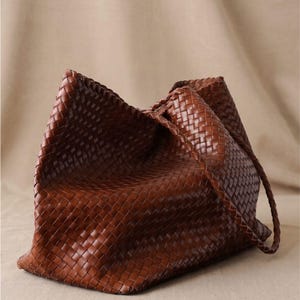 May include: A dark brown tote bag featuring an intricate woven pattern across its entire surface. The bag has a structured, rectangular base and includes two thin, braided shoulder straps attached to the top edges of the main body.