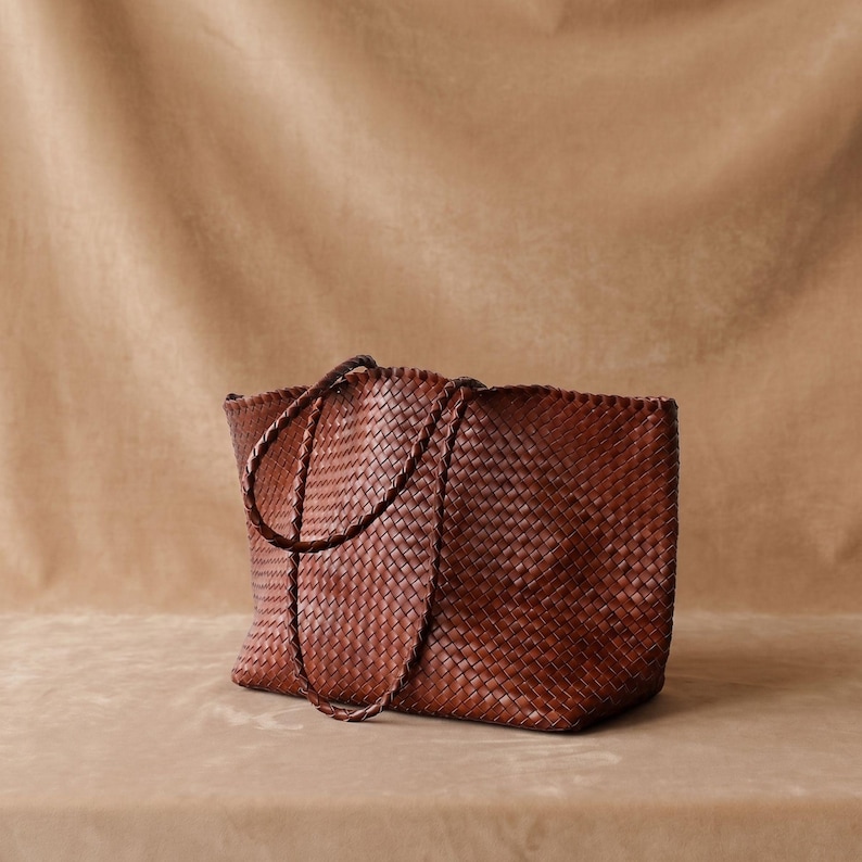 May include: A dark brown tote bag constructed from woven leather strips. The bag features a rectangular base, an open top, and two thin, braided leather shoulder straps that are attached to the upper edges of the bag body.