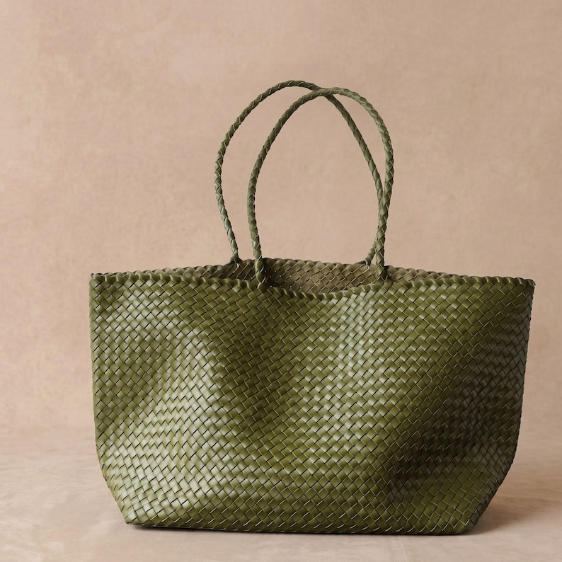 May include: An olive green tote bag featuring a woven construction with a consistent grid pattern throughout the body. The bag includes two thin, braided handles attached to the top edge, providing a structured yet flexible design for carrying.