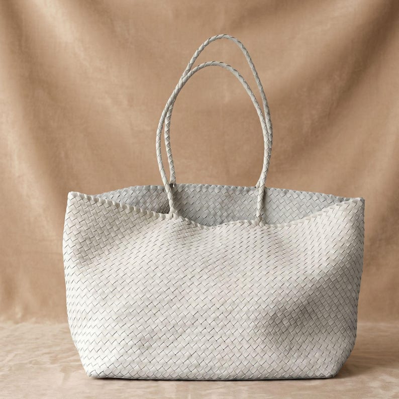 May include: A light grey tote bag featuring an intricate woven pattern throughout the entire exterior. The bag includes two long, slender braided handles attached to the top edge, providing a consistent texture and a structured open-top design.