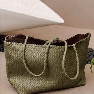 May include: An olive green tote bag featuring a woven exterior pattern and two matching braided shoulder straps. The interior of the bag is lined with a dark brown material, and the overall construction displays a consistent, uniform grid-like texture.