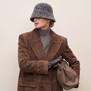 May include: A charcoal gray textured bucket hat worn with a brown suede blazer, a gray mock neck top, dark brown leather gloves, and a taupe leather handbag. The ensemble features structured lapels, button details on the sleeves, and pockets.