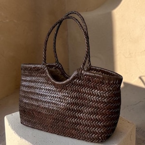 May include: A dark brown woven leather tote bag featuring a tight herringbone pattern throughout the body. The bag has two thin, braided leather handles attached to the top edge, which curves downward in the center to form a U-shaped opening.