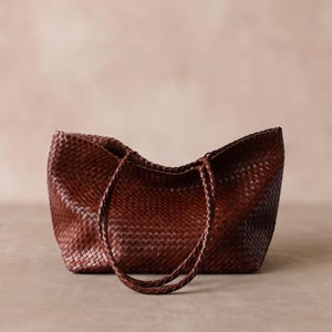 May include: A dark brown tote bag featuring a woven texture throughout the entire exterior. The bag has two thin, braided shoulder straps attached to the top edge, creating a structured yet flexible silhouette with a wide, open main compartment.