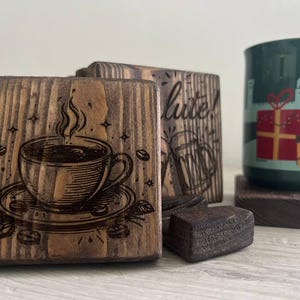 Può includere: Sottobicchieri in legno a tema caffè. Uno presenta un'incisione dettagliata di una tazza di caffè con vapore, chicchi e foglie. Una tazza verde con un disegno regalo poggia su un sottobicchiere. I sottobicchieri sono in legno scuro.