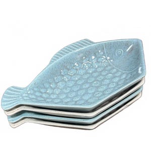 May include: Four light blue ceramic plates, each shaped like a fish. The plates have a textured, speckled surface. They are stacked, with the top plate showing the fish's head and tail. Each plate is approximately 25 cm long.