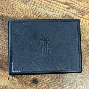 Puede incluir: Un estuche rectangular de plástico negro para naipes con una superficie texturizada. El centro presenta un logotipo en relieve con un icono de corona sobre las letras KEM. El texto KEM PLASTIC PLAYING CARDS está grabado en la parte inferior.
