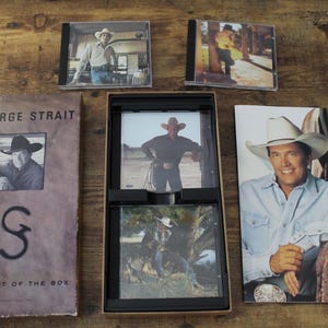 Könnte beinhalten: Eine George Strait Strait Out of the Box Musiksammlung mit einer braunen Pappschachtel mit dem Namen und Logo des Künstlers, zwei separaten CD-Hüllen, einem Box-Einsatz mit zwei weiteren CDs und einem hohen Booklet mit einem Künstlerporträt.