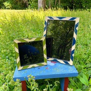 May include: Two decorative mirrors with patterned frames rest on a blue bench in a garden setting. One mirror has a green and cream zigzag pattern, while the other features a blue and cream geometric design. The mirrors reflect the surrounding greenery.