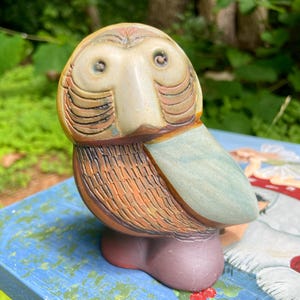 May include: A ceramic owl figurine featuring a rounded head with two small eyes, a smooth beige face, and a textured body with brown and orange plumage. The wings are a muted light blue, and the base is a solid, rounded dark purple shape.