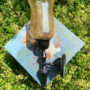 May include: A wall-mounted sconce with a crackled amber glass shade and a black metal base. The sconce is mounted on a blue square with a fairy tale illustration. The sconce is surrounded by green foliage.