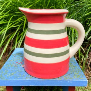 May include: A ceramic pitcher featuring a repeating horizontal striped pattern in vibrant red, off-white, and muted olive green. The vessel has a wide, curved handle on the right side and a flared pouring spout at the top rim with a smooth finish.