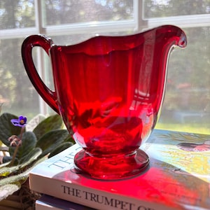 May include: A translucent ruby red glass creamer pitcher featuring a scalloped rim, a curved handle, and a circular pedestal base. The pitcher has a smooth, glossy finish and a distinct pouring spout, catching light to create deep red reflections.