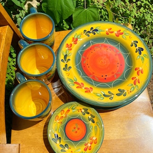 May include: A ceramic dinnerware set featuring three mugs, one large dinner plate, and one small plate. Each piece has a bright yellow interior, a central orange circle, and a border decorated with green leaves and red floral accents.