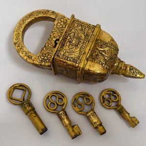 Puede incluir: Un candado de metal dorado con intrincados grabados de flores y hojas en su superficie, acompañado de cuatro llaves a juego con cabezas circulares distintivas, cada una con patrones detallados en los vástagos y los arcos.