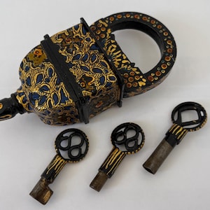 Puede incluir: Un candado antiguo ornamentado con un asa a juego y tres llaves. El candado y el asa son negros con patrones florales dorados y azules. Las llaves tienen diseños decorativos negros y dorados.
