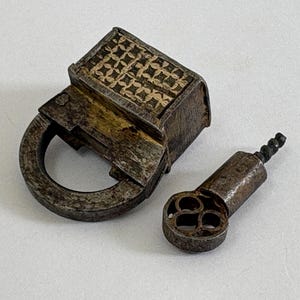 May include: A weathered metal padlock featuring a rectangular body with an intricate, repeating geometric pattern on its top surface and a circular shackle. Beside the padlock lies a matching metal key with a decorative, openwork circular bow.