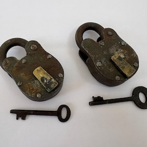 Puede incluir: Dos candados de hierro desgastados con cubiertas de latón para la cerradura y llaves maestras a juego. Los candados presentan oxidación visible, remaches y texto estampado en las superficies metálicas. Cada candado tiene un arco redondeado.