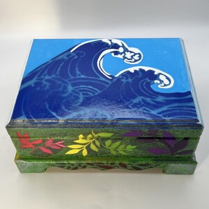 Op de afbeelding: Een rechthoekige houten doos met een beschilderd deksel met een blauwe oceaangolf en witte schuimkoppen. De zijkanten van de doos zijn groen geschilderd en versierd met een patroon van kleine, kleurrijke blaadjes in rood, geel en paars.